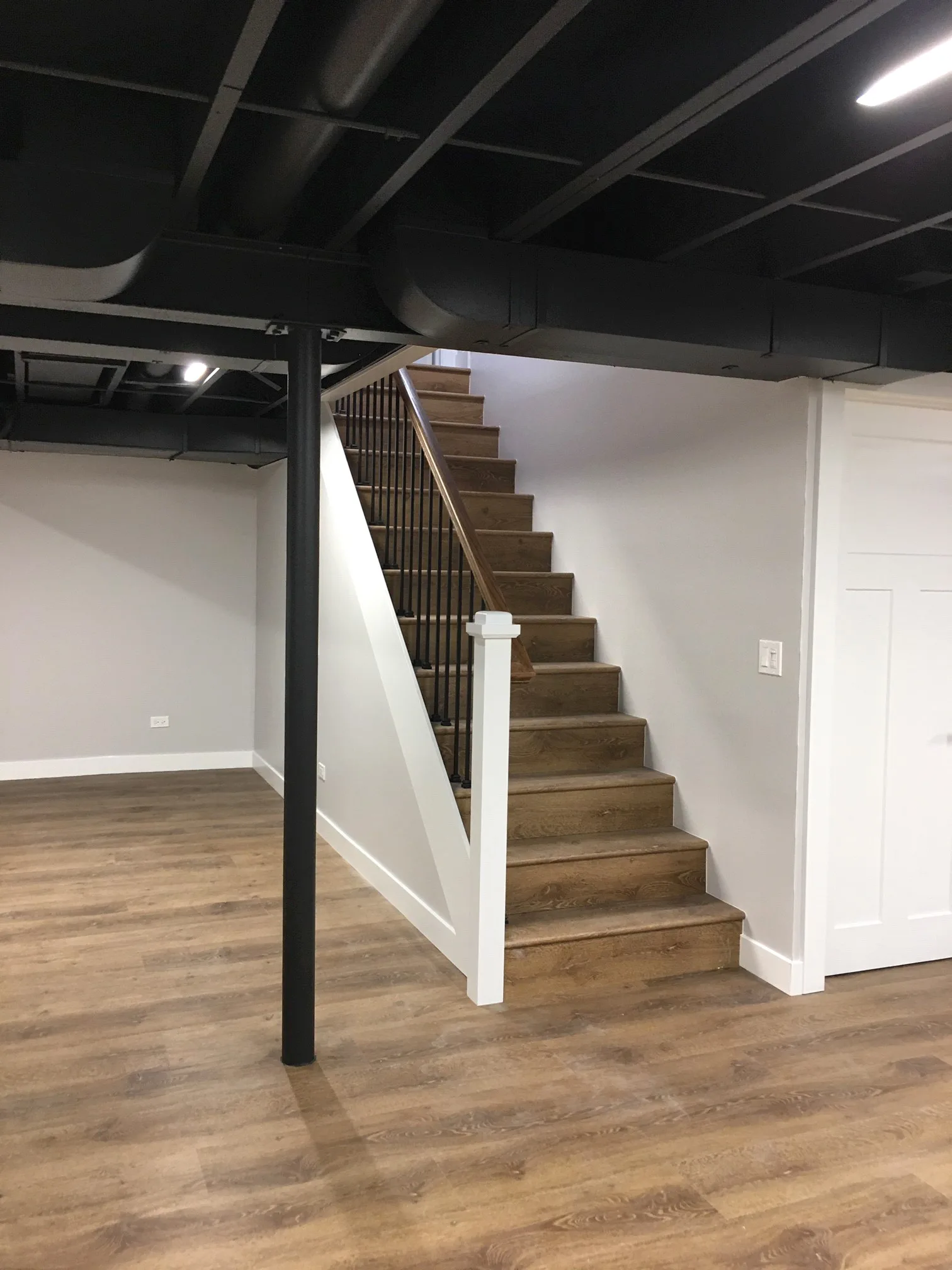 Lake Forest basement buildout by Delta: exposed black-painted ceiling joists, refinished oak staircase with iron spindle railing, light gray walls, wood-look plank flooring