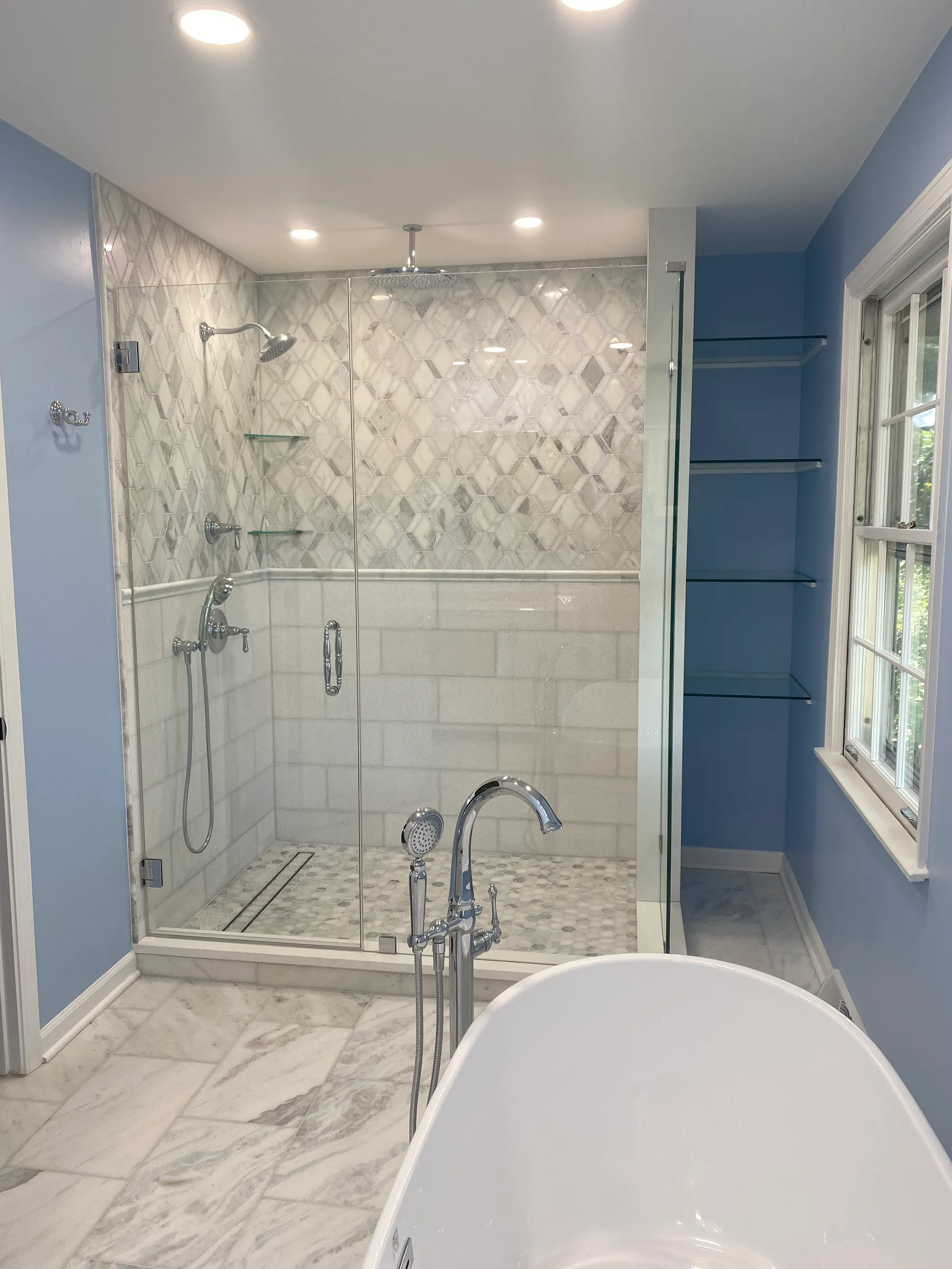 Lake Bluff primary bathroom by Delta: frameless glass walk-in shower with carrara marble diamond-mosaic feature wall, ceiling rainhead, freestanding soaker tub, chrome floor-mount filler