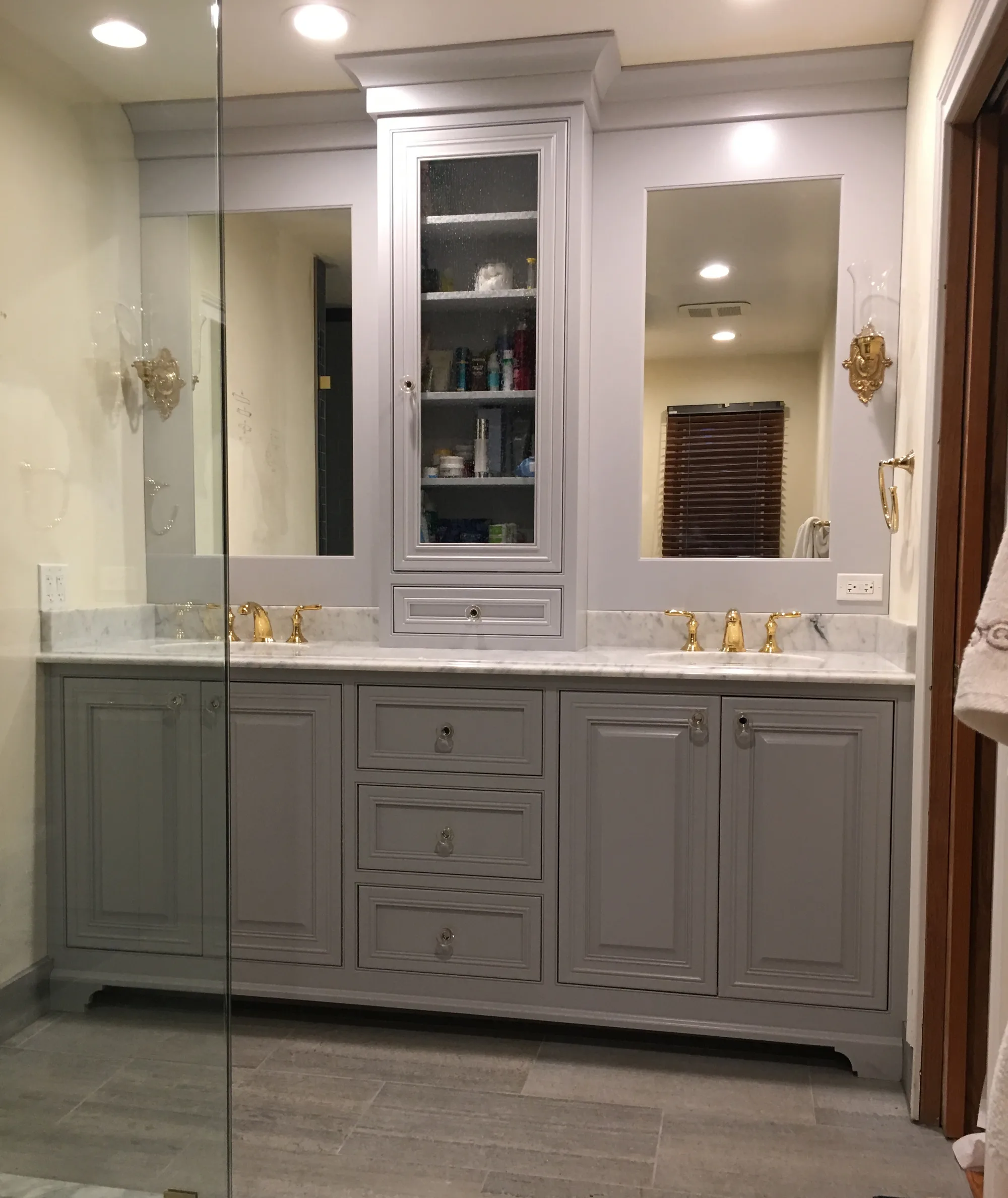 Lake Forest primary bathroom by Delta: gray raised-panel double vanity with carrara marble counters, polished brass faucets, and frameless glass walk-in shower