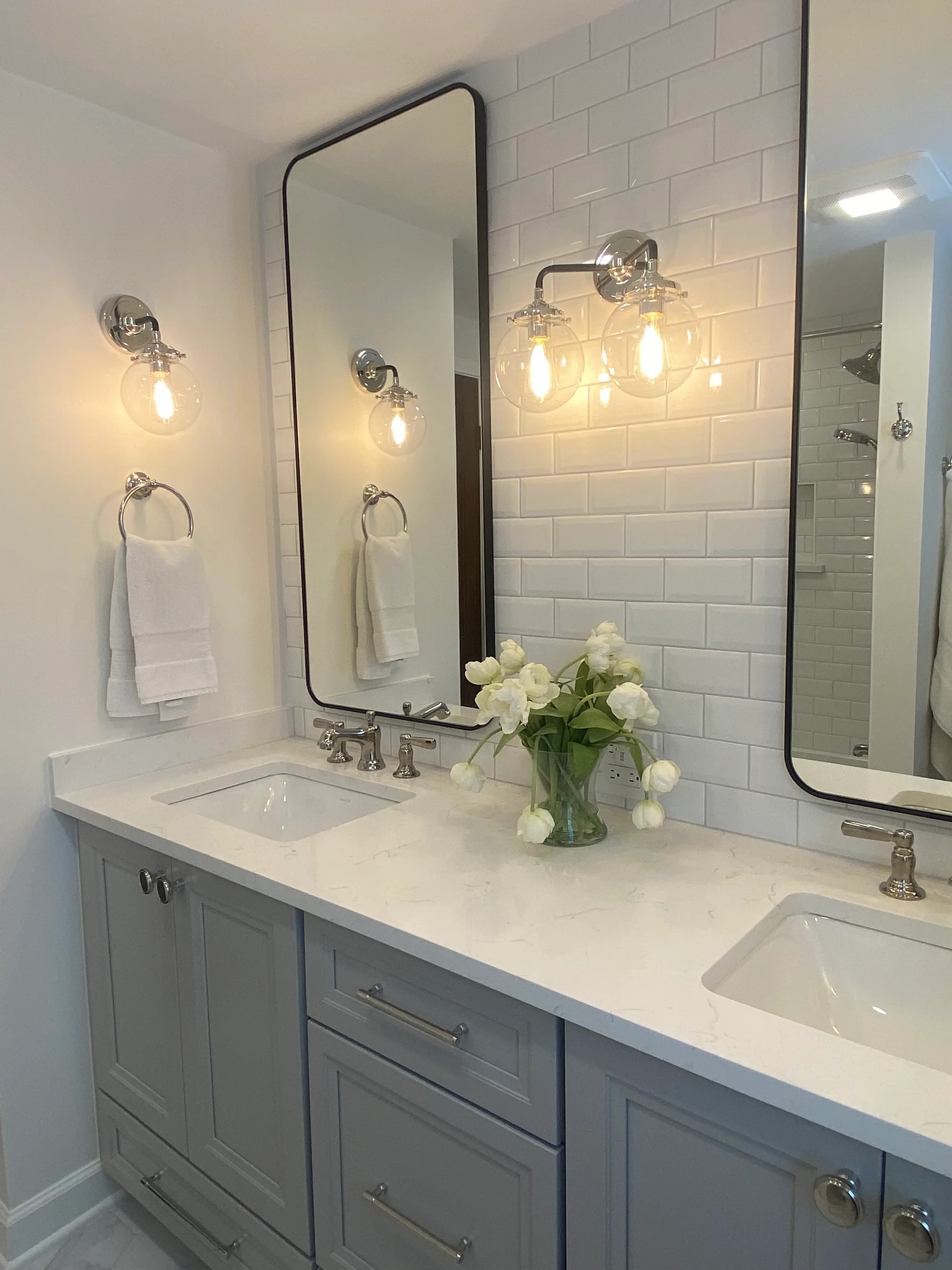 Northbrook bathroom by Delta: gray inset shaker double vanity, white quartz counter, white subway backsplash, black-framed pivot mirrors, cage globe sconces