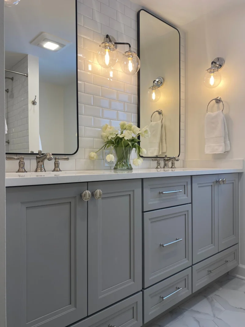 Gray double vanity with quartz top, beveled subway tile, black-framed mirrors and glass globe sconces