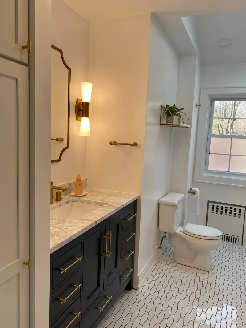 Bathroom with a navy vanity, marble top, brass hardware and sconce, and white elongated hexagon floor tile