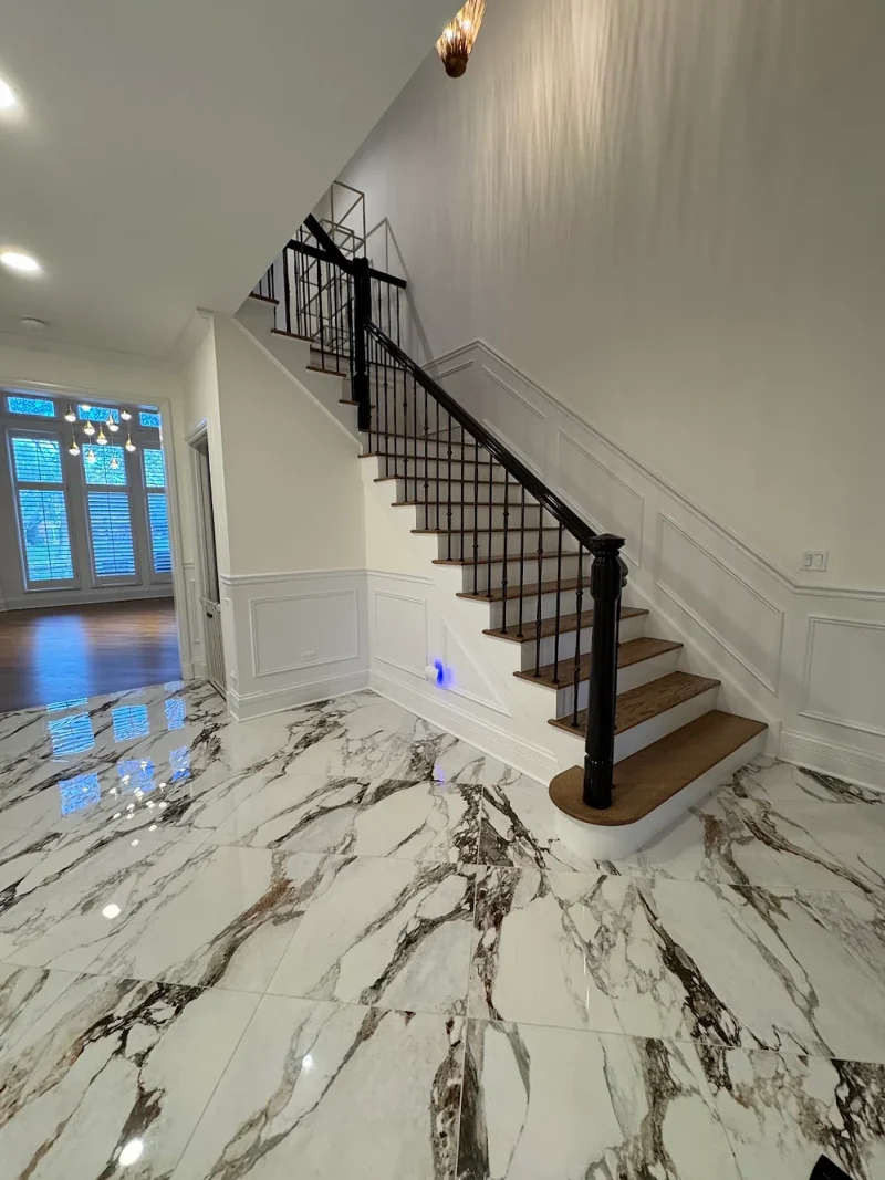 Foyer staircase with black railing and newel post, oak treads, raised-panel wainscoting and large marble-look floor tile