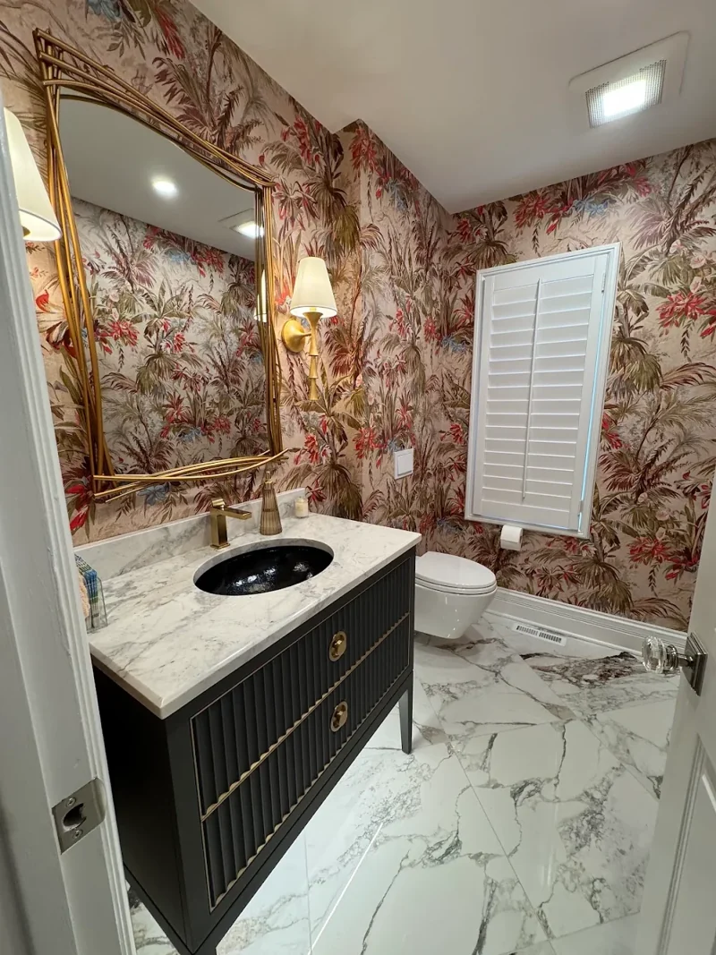 Powder room with tropical wallpaper, gold mirror and sconce, black fluted vanity with marble top and marble-look floor tile