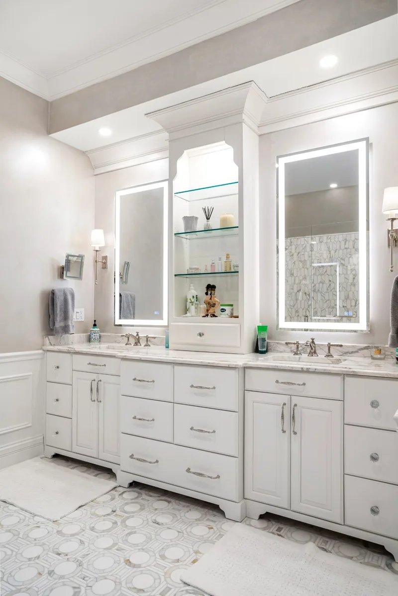 White double vanity with marble counter, lighted mirrors, wall sconces and a center tower with glass shelves