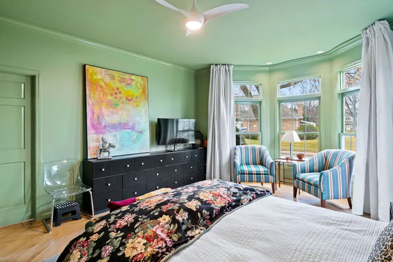 Green primary bedroom with a black dresser, bay windows and two striped armchairs