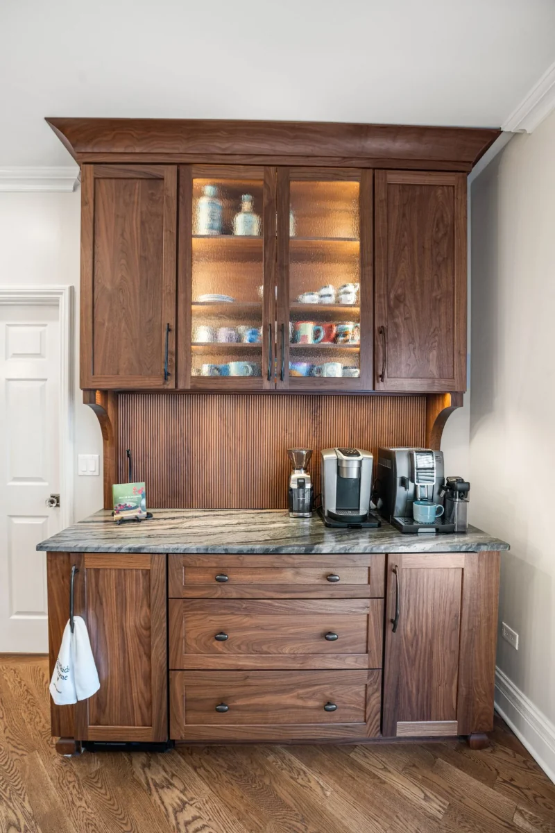 Walnut coffee bar with lit glass-front cabinets, fluted wood backsplash and a leathered stone counter
