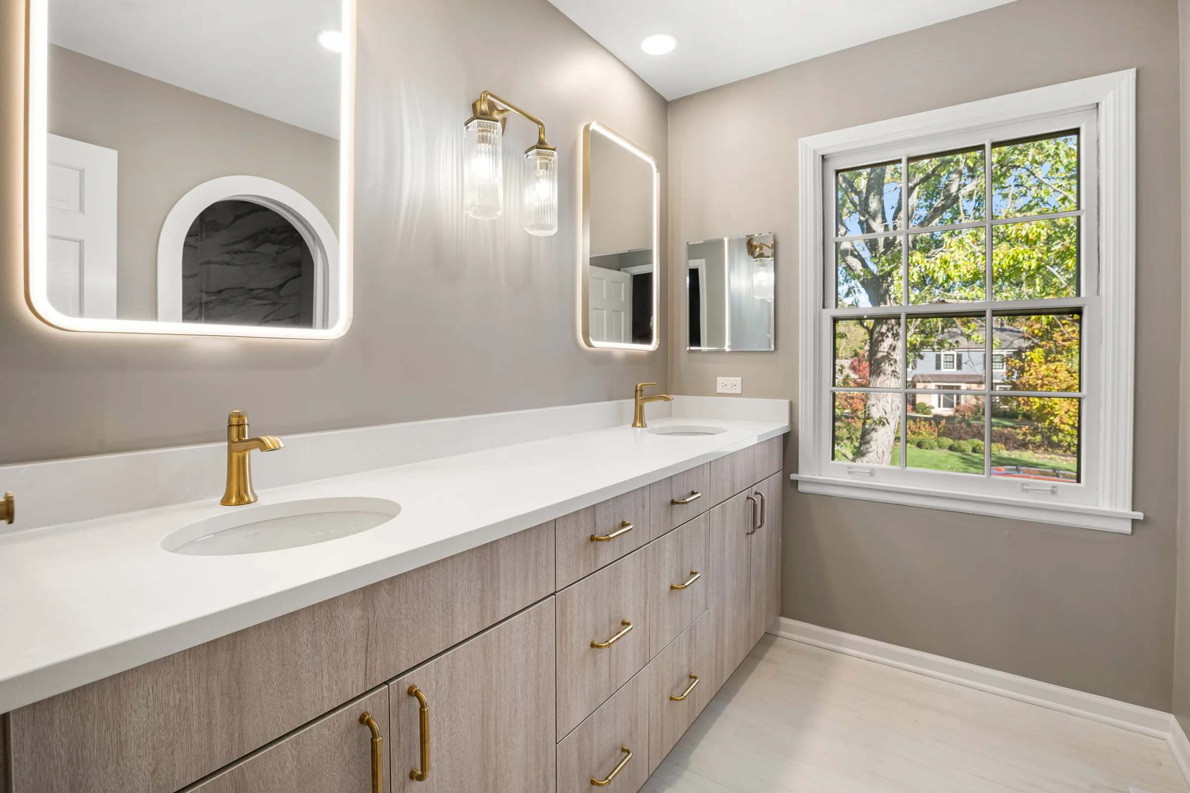 Lake Forest primary bathroom: double vanity with brushed brass fixtures and backlit arched mirrors