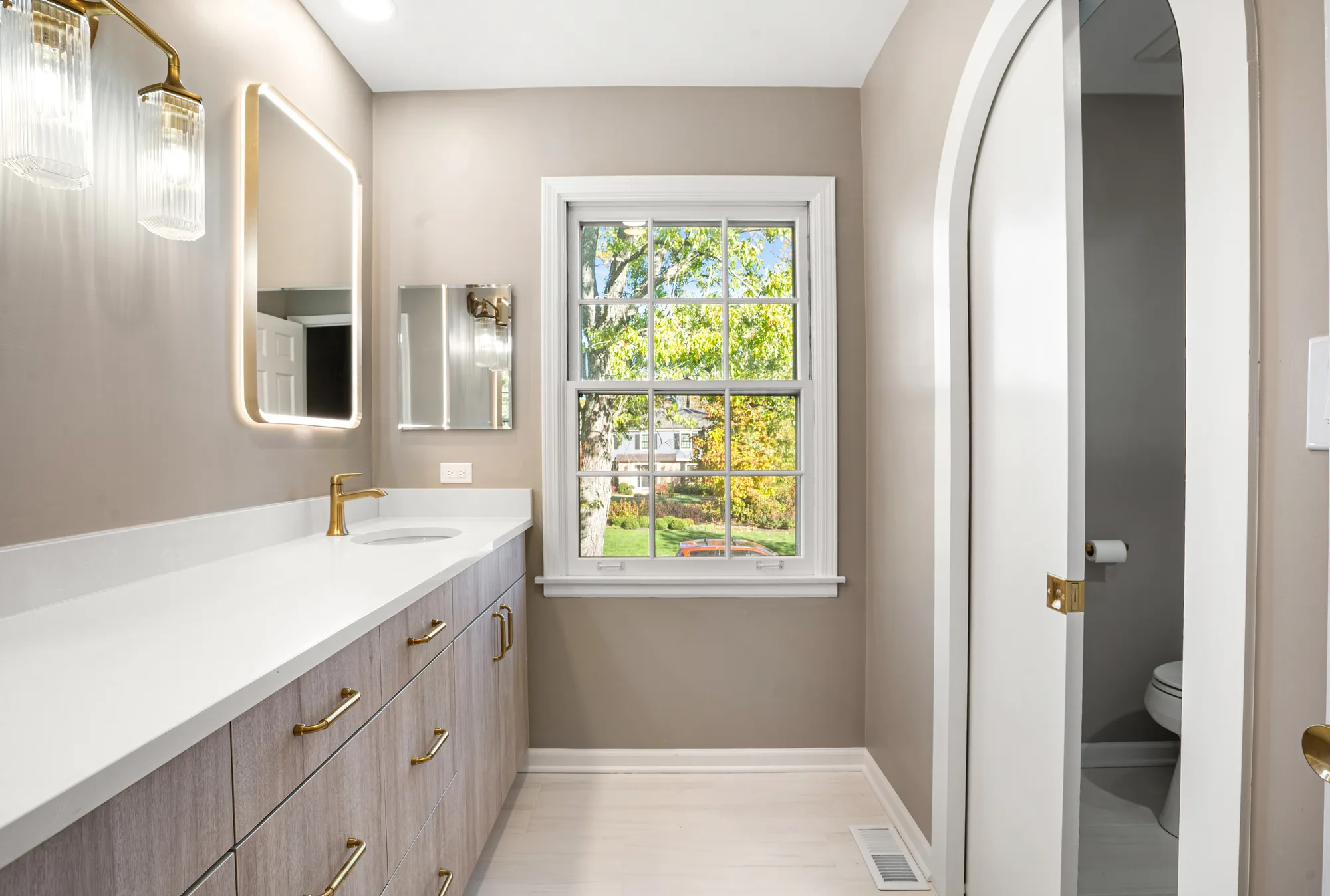 Wilson Road bathroom Lake Forest: light-wood flat-panel vanity with brushed brass pulls, white quartz counter, backlit LED mirror, fluted glass sconces